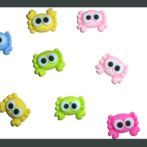 May include: A collection of colorful, cartoon-like crab-shaped embellishments. The crabs are in various colors, including blue, yellow, green, pink, and purple, each with large, googly eyes. These decorative elements are likely used for crafts or embellishments.