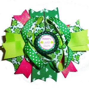 Puede incluir: Lazo para el pelo verde y rosa con un patrón de trébol verde y un dije redondo que dice "Mommy's Lucky Charm".