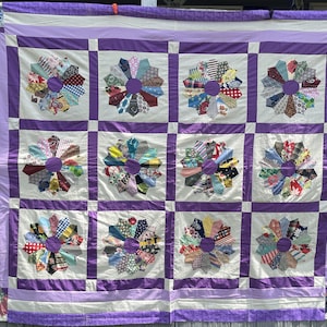 Op de afbeelding: Een quilt met een raster van twaalf vierkante blokken, elk met een cirkelvormig, bloemachtig patroon gemaakt van diverse stoffen met verschillende prints. De blokken worden gescheiden door paarse stoffen stroken.