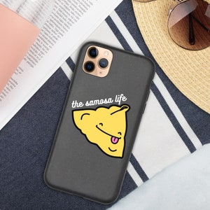 Puede incluir: Una funda de teléfono negra con una superficie texturizada. La funda tiene un gráfico blanco de un samosa amarillo con una lengua rosa y el texto "the samosa life" en blanco.