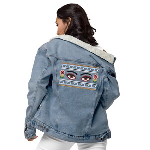 May include: A light blue denim jacket with a sherpa lining and a colorful embroidered patch on the back. The patch features two stylized eyes with red, yellow, and blue accents, surrounded by a geometric pattern.