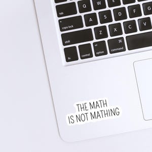 May include: A white sticker with black text that reads "THE MATH IS NOT MATHING" is placed on a laptop. The laptop keyboard is visible, with black keys and white lettering. The sticker is rectangular with rounded corners.