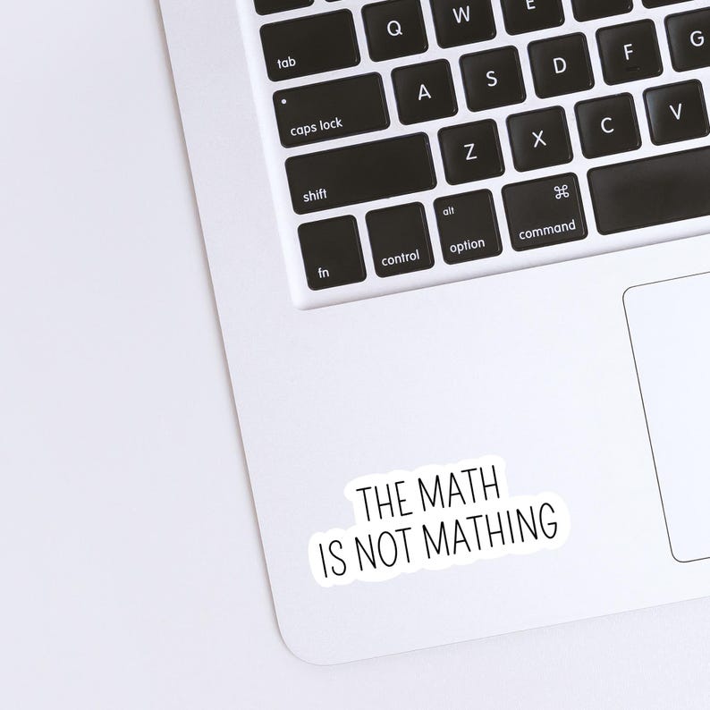 May include: A white sticker with black text that reads "THE MATH IS NOT MATHING" is placed on a laptop. The laptop keyboard is visible, with black keys and white lettering. The sticker is rectangular with rounded corners.