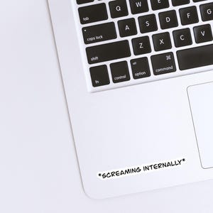 Op de afbeelding: Een witte sticker met de tekst "*SCREAMING INTERNALLY*" in het zwart, geplaatst op een laptop. Het toetsenbord van de laptop is zichtbaar, met zwarte toetsen en witte letters. De sticker is rechthoekig en heeft een zwarte rand.