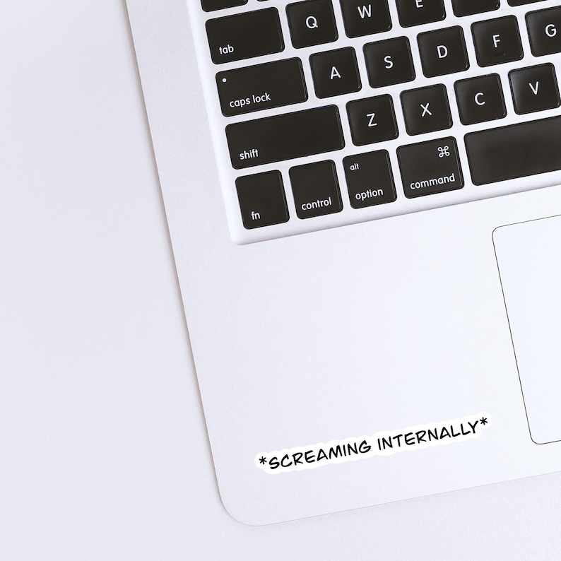 Op de afbeelding: Een witte sticker met de tekst "*SCREAMING INTERNALLY*" in het zwart, geplaatst op een laptop. Het toetsenbord van de laptop is zichtbaar, met zwarte toetsen en witte letters. De sticker is rechthoekig en heeft een zwarte rand.
