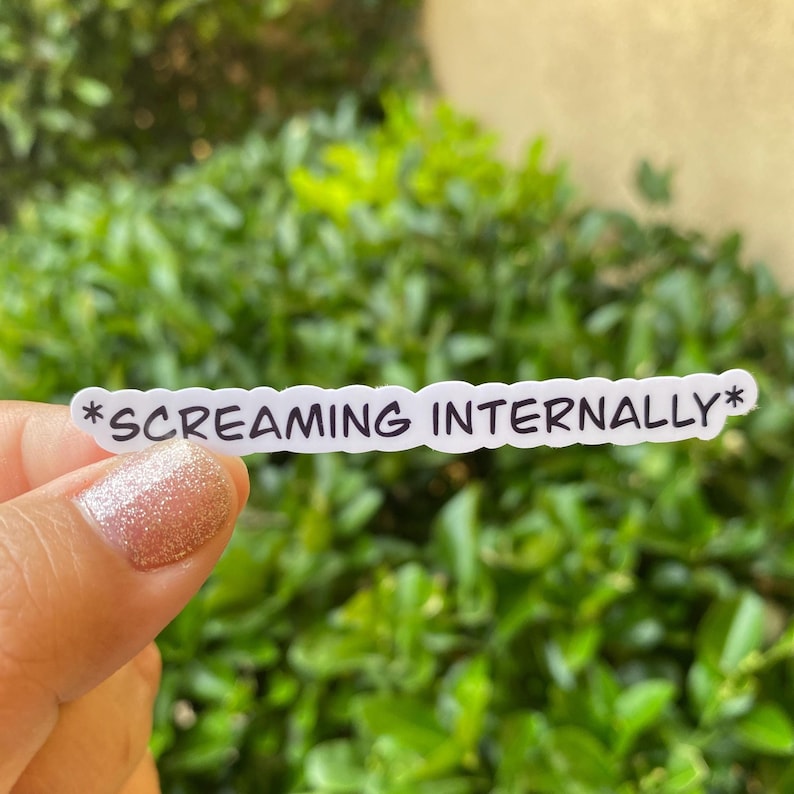 Op de afbeelding: Een witte sticker met de tekst "*SCREAMING INTERNALLY*" in zwart, tegen een achtergrond van groene bladeren. De sticker heeft een golvende rand. De tekst is in een schreefloze lettertype.