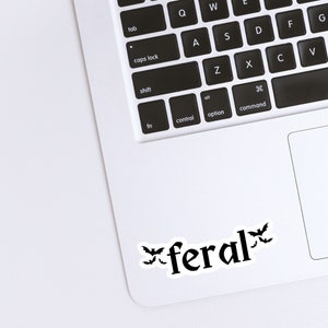 May include: A white laptop computer with a black keyboard and a black trackpad. The word "feral" is written in black with a stylized font and three black bats are on either side of the word.