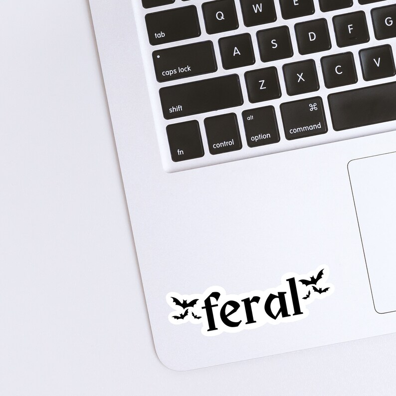 May include: A white laptop computer with a black keyboard and a black trackpad. The word "feral" is written in black with a stylized font and three black bats are on either side of the word.