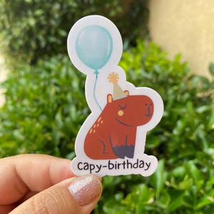 May include: A sticker featuring a cartoon capybara wearing a party hat and holding a blue balloon. The sticker says "capy-birthday" in white text.
