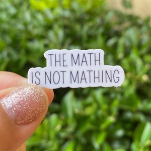 Könnte beinhalten: Ein weißer Aufkleber mit dem Text "THE MATH IS NOT MATHING" in Schwarz, vor einem verschwommenen grünen Hintergrund. Der Aufkleber hat eine abgerundete rechteckige Form.