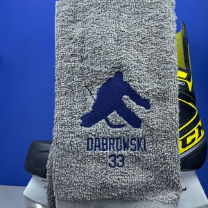 May include: A gray hockey towel with a dark blue embroidered hockey player silhouette, the name "DABROWSKI" and the number "33". A hockey skate is visible in the background.