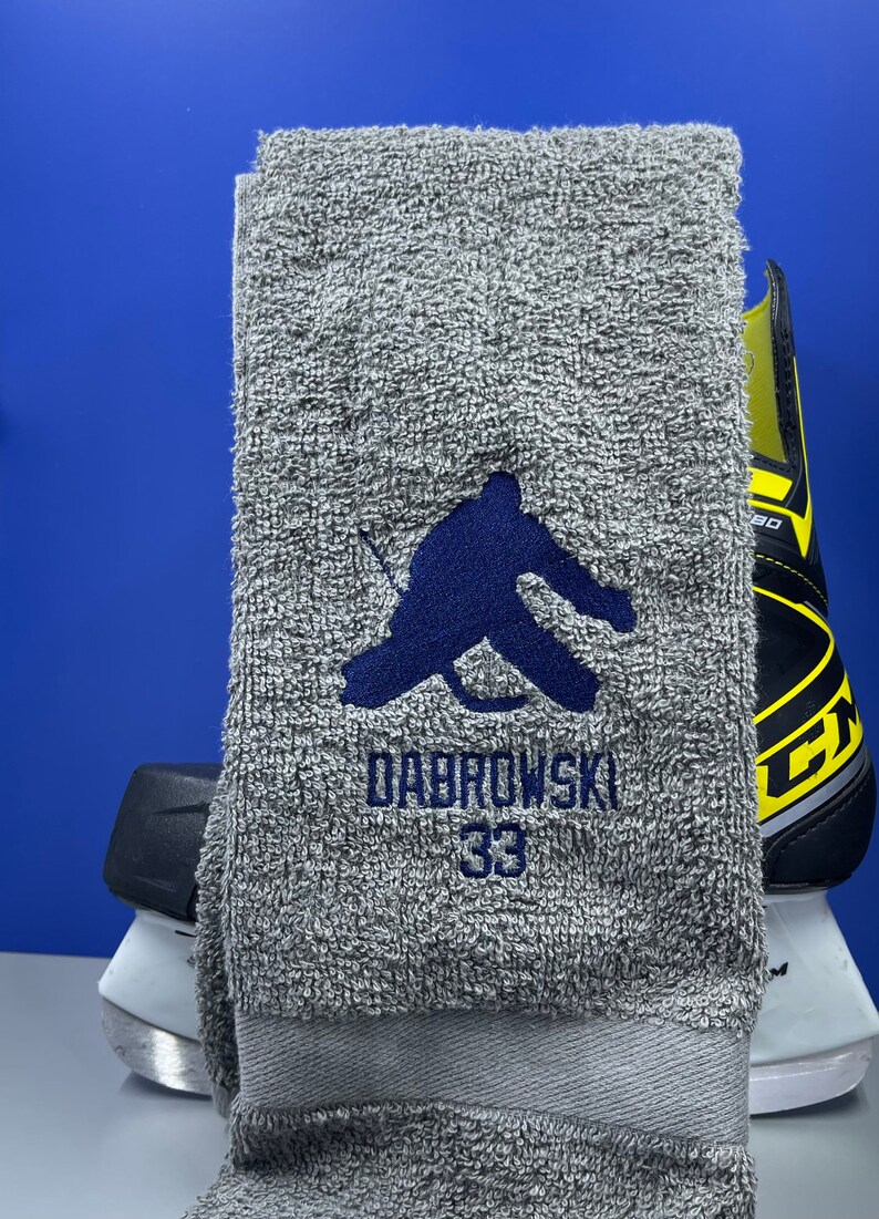 May include: A gray hockey towel with a dark blue embroidered hockey player silhouette, the name "DABROWSKI" and the number "33". A hockey skate is visible in the background.