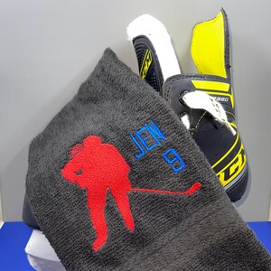 May include: A black hockey skate with yellow accents and white laces is next to a dark gray towel. The towel has a red hockey player silhouette and the name "JEN 9" embroidered in blue.