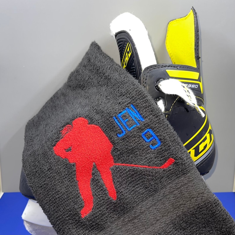 May include: A black hockey skate with yellow accents and white laces is next to a dark gray towel. The towel has a red hockey player silhouette and the name "JEN 9" embroidered in blue.