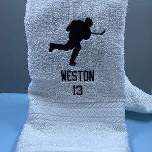 May include: White hockey towel with a black silhouette of a hockey player in action. The name "WESTON" and the number "13" are embroidered in black below the image. The towel is folded and has a textured surface.