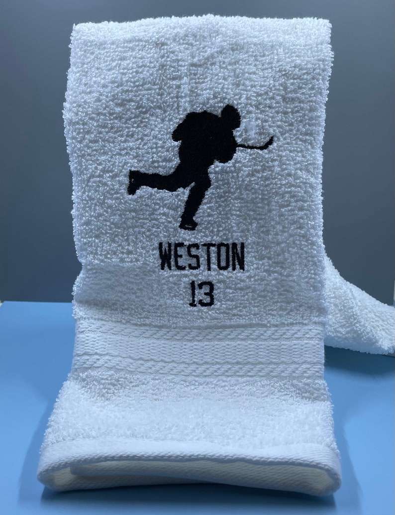 May include: White hockey towel with a black silhouette of a hockey player in action. The name "WESTON" and the number "13" are embroidered in black below the image. The towel is folded and has a textured surface.