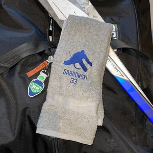 May include: A gray hockey towel embroidered with a blue hockey player silhouette, the name "DABROWSKI" and the number "33". The towel is folded and rests on a black bag, with hockey sticks visible in the background. A hockey mask keychain hangs from the bag.