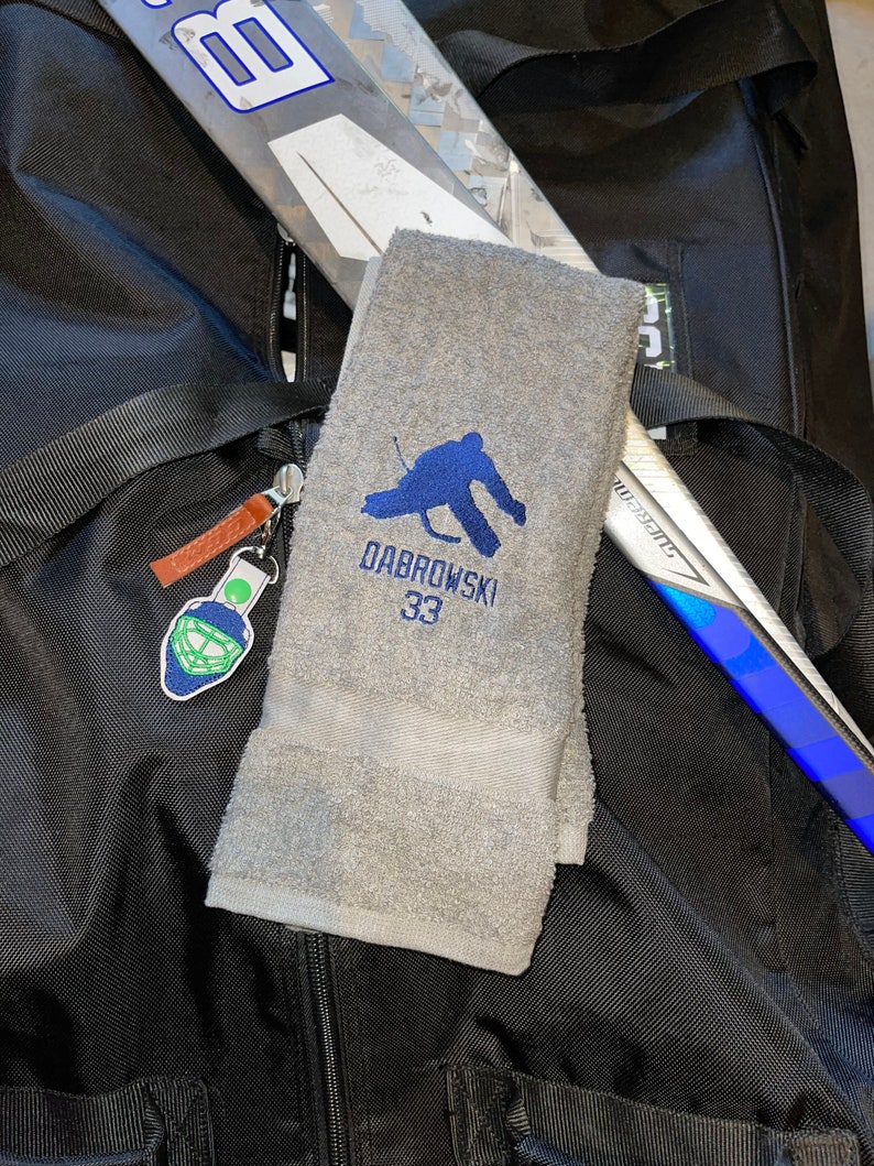 May include: A gray hockey towel embroidered with a blue hockey player silhouette, the name "DABROWSKI" and the number "33". The towel is folded and rests on a black bag, with hockey sticks visible in the background. A hockey mask keychain hangs from the bag.