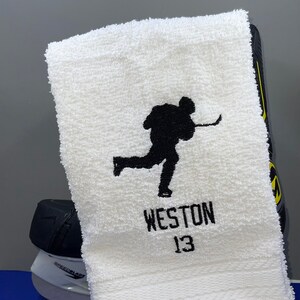 May include: White hockey towel with a black silhouette of a hockey player in action. The name "WESTON" and the number "13" are embroidered in black below the player. A hockey skate is visible in the background.