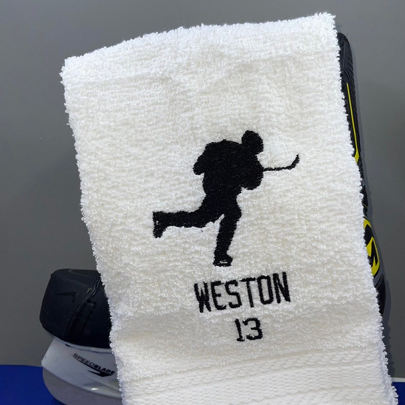 May include: White hockey towel with a black silhouette of a hockey player in action. The name "WESTON" and the number "13" are embroidered in black below the player. A hockey skate is visible in the background.