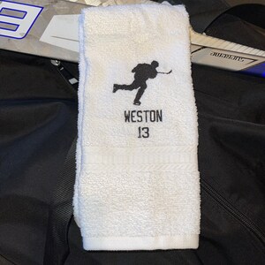 May include: White hockey towel with a black embroidered silhouette of a hockey player. The name "WESTON" and the number "13" are embroidered below the player. A hockey stick and a black bag are in the background.