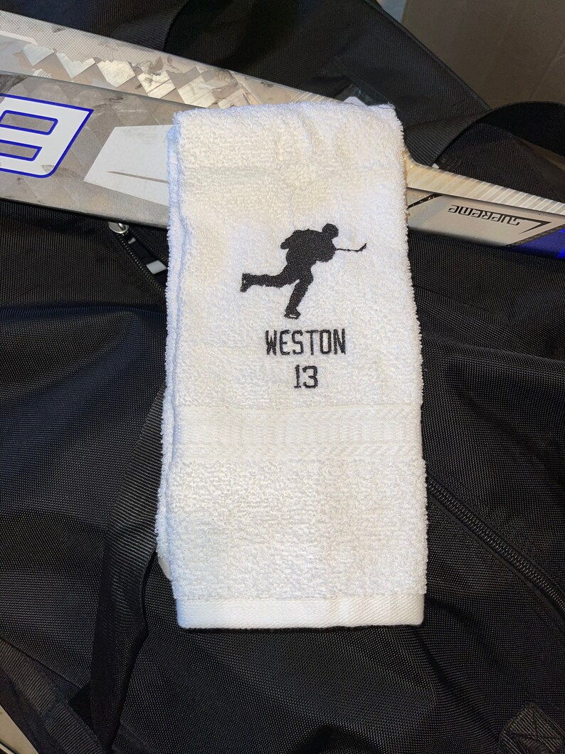 May include: White hockey towel with a black embroidered silhouette of a hockey player. The name "WESTON" and the number "13" are embroidered below the player. A hockey stick and a black bag are in the background.