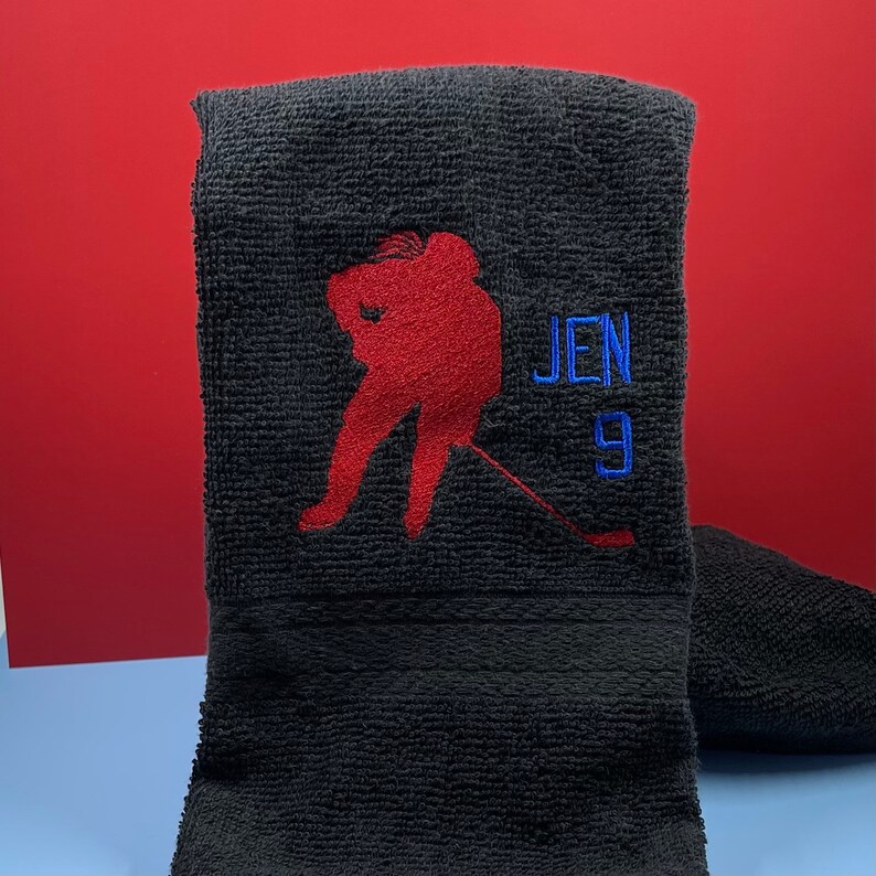 May include: Black hand towel with a red embroidered hockey player silhouette. The name "JEN" and the number "9" are embroidered in blue. The towel is folded and set against a red background.