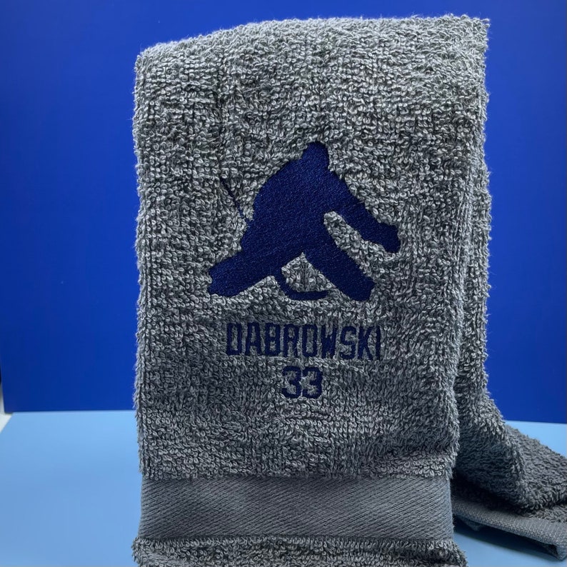 May include: A gray towel with a dark blue embroidered design and the name "DABROWSKI" and the number "33". The towel is folded and has a textured appearance.