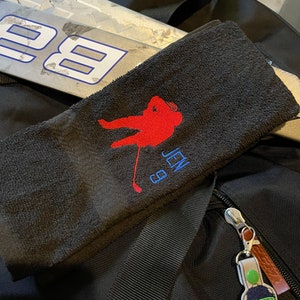 May include: Black hockey towel with a red hockey player silhouette, the name "JEN" and the number "9" embroidered in blue. A hockey stick is visible in the background. A hockey goalie mask keychain is attached to a black bag.