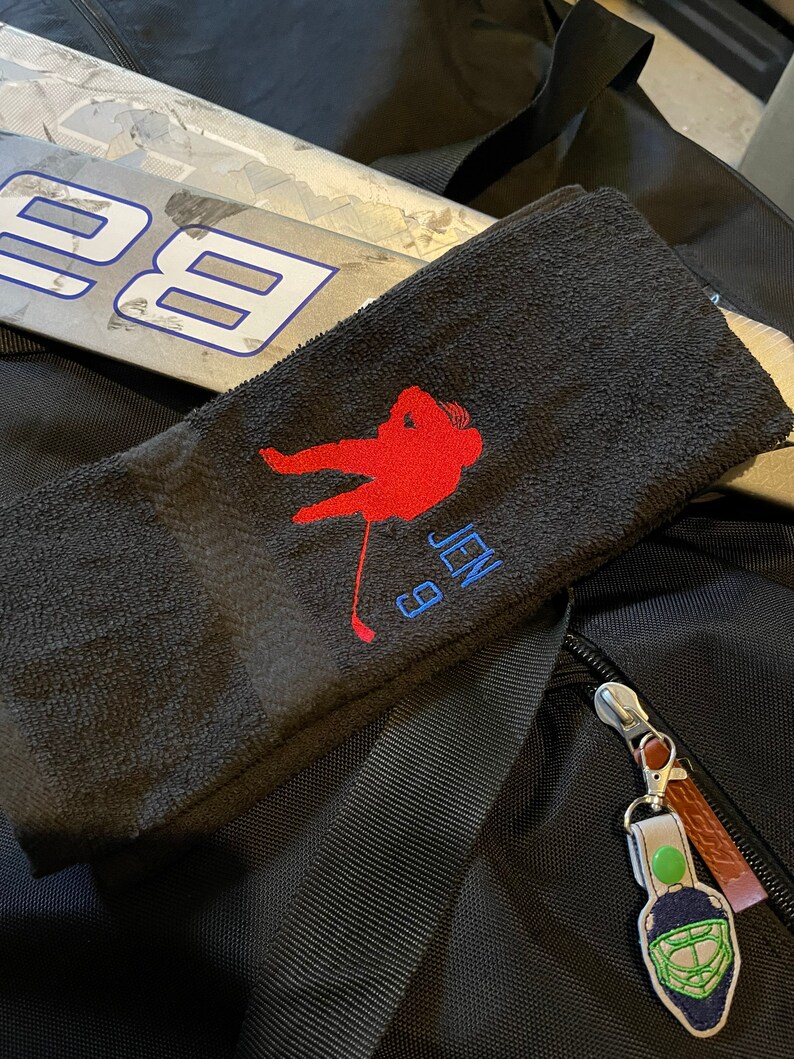 May include: Black hockey towel with a red hockey player silhouette, the name "JEN" and the number "9" embroidered in blue. A hockey stick is visible in the background. A hockey goalie mask keychain is attached to a black bag.