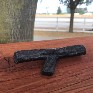 Könnte beinhalten: Ein schwarzer, T-förmiger Türstopper oder Möbelstopper aus Metall, der auf einer Holzoberfläche liegt.