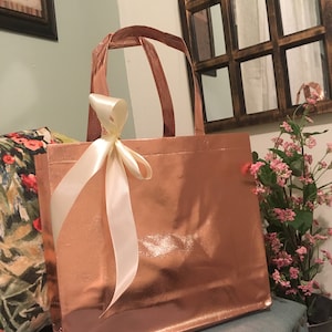 May include: A rose gold tote bag with matching handles and a cream-colored ribbon tied in a bow. The bag is sitting on a blue surface, with a floral pillow and a decorative mirror in the background.