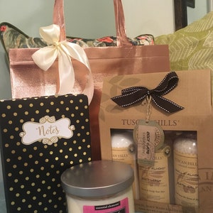 May include: A gift set featuring a rose gold tote bag with a cream ribbon, a black notebook with gold polka dots, a candle, and a Tuscan Hills body care set. The candle label reads "Raspberry & Geranium".