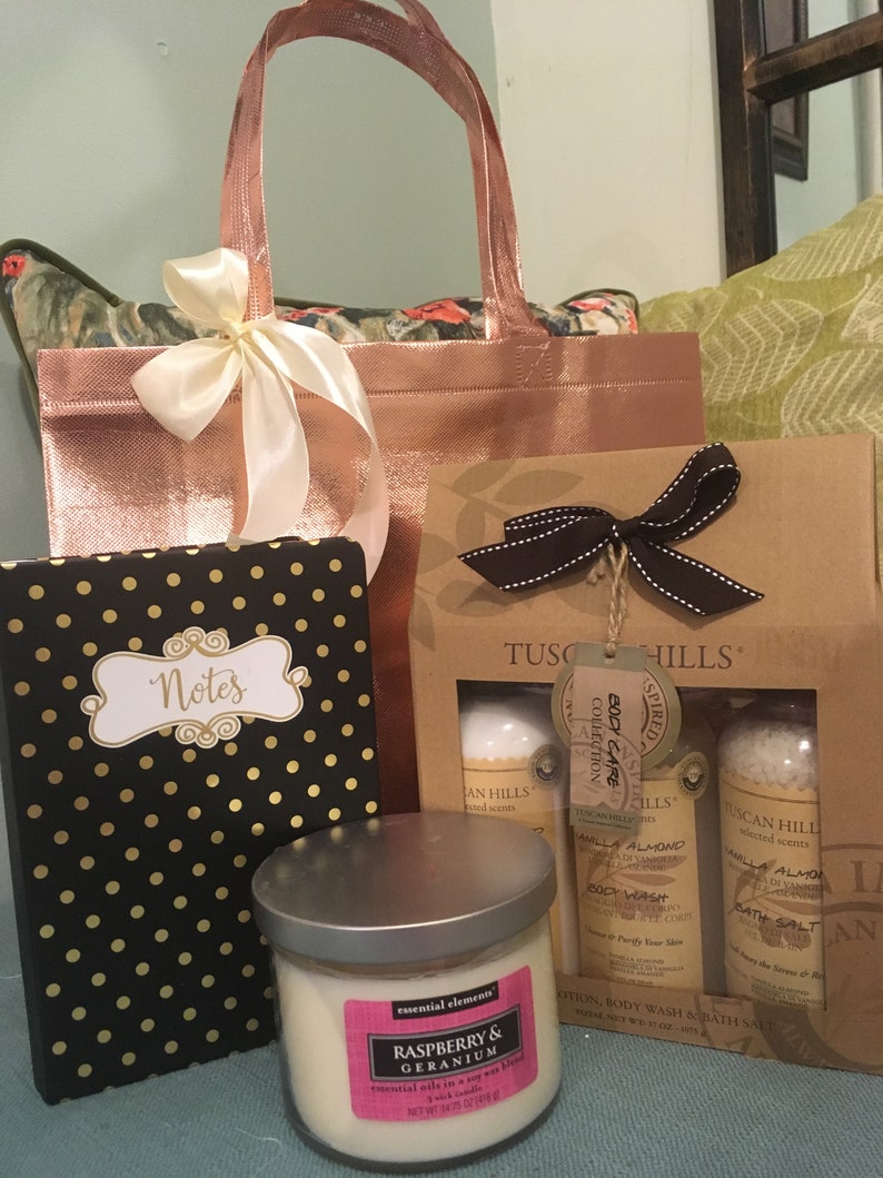 May include: A gift set featuring a rose gold tote bag with a cream ribbon, a black notebook with gold polka dots, a candle, and a Tuscan Hills body care set. The candle label reads "Raspberry & Geranium".