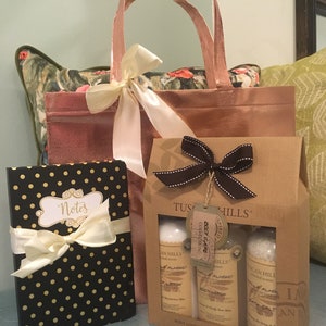 May include: A gift set featuring a rose gold tote bag tied with a cream ribbon, a black notebook with gold polka dots and a cream bow, and a Tuscan Hills body care set in a brown box. The body care set includes lotion, body wash, and bath salt.