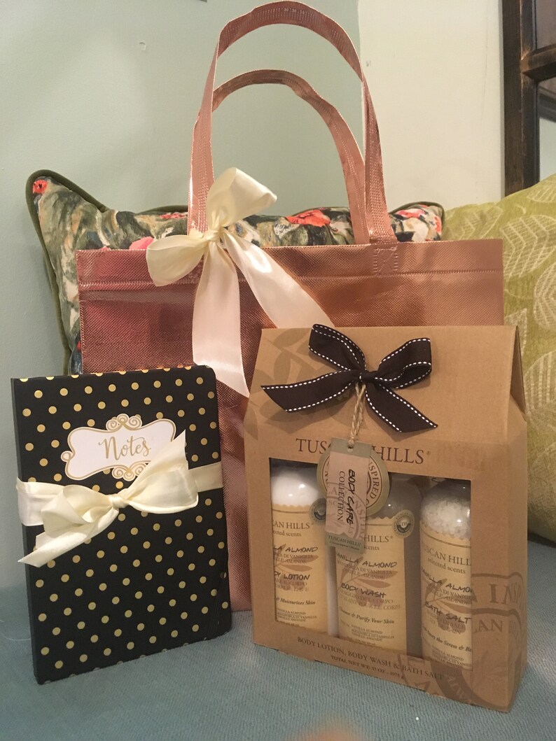 May include: A gift set featuring a rose gold tote bag tied with a cream ribbon, a black notebook with gold polka dots and a cream bow, and a Tuscan Hills body care set in a brown box. The body care set includes lotion, body wash, and bath salt.