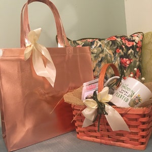 May include: A rose gold tote bag and a small orange woven basket with a handle. The basket contains a mug with the text "Bloom where you are planted". Both are decorated with cream-colored ribbons.