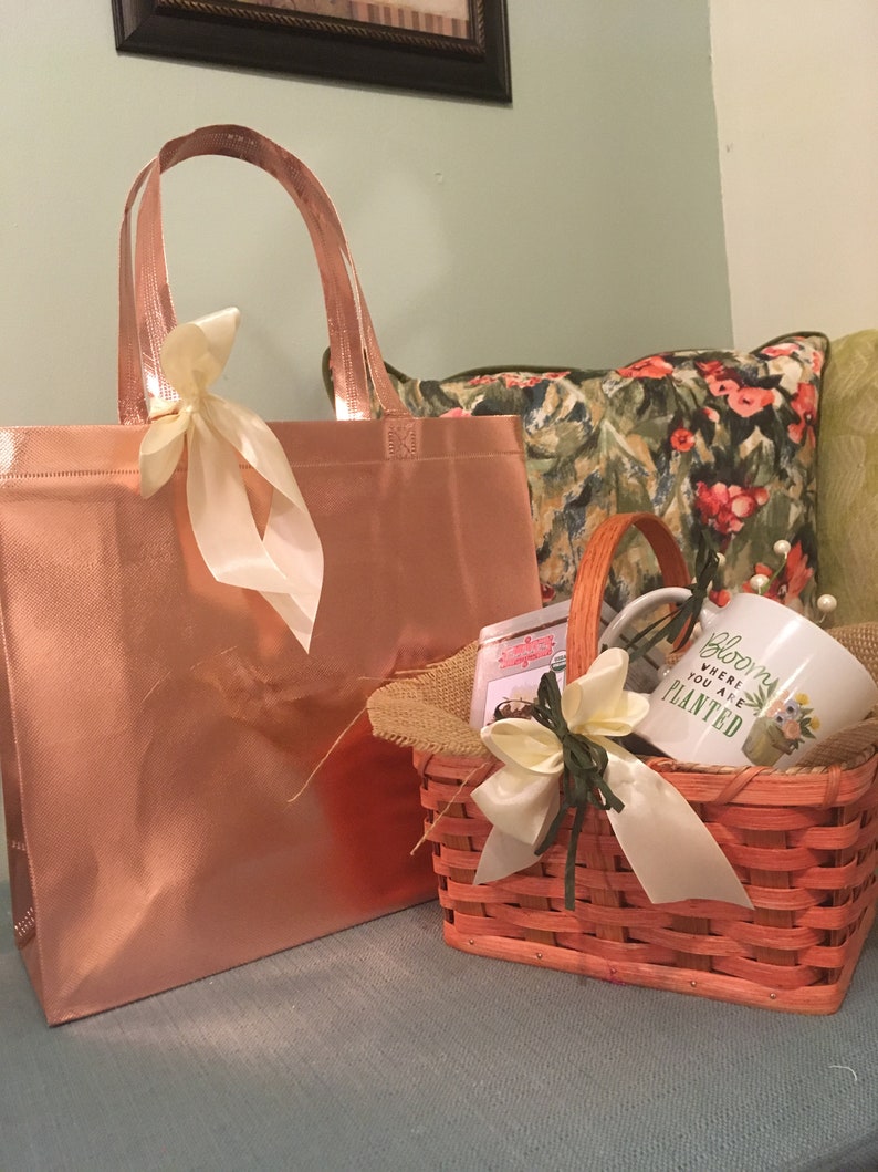 May include: A rose gold tote bag and a small orange woven basket with a handle. The basket contains a mug with the text "Bloom where you are planted". Both are decorated with cream-colored ribbons.