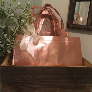 May include: Rose gold tote bags with matching handles and a white ribbon bow, nestled inside a rustic wooden box. The bags have a shiny, metallic finish. White flowers and a mirror are in the background.