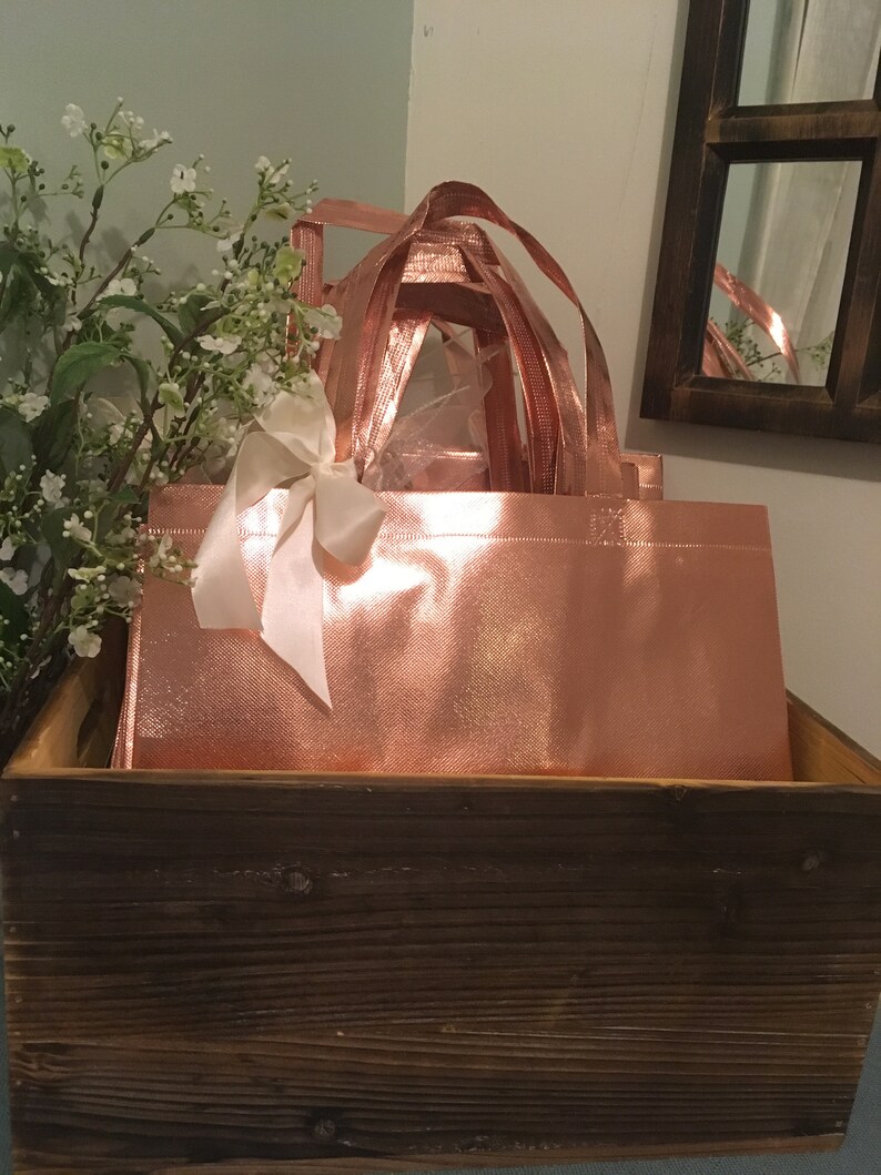 May include: Rose gold tote bags with matching handles and a white ribbon bow, nestled inside a rustic wooden box. The bags have a shiny, metallic finish. White flowers and a mirror are in the background.