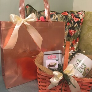 May include: A gift basket with a rose gold tote bag and a small orange woven basket. The basket contains a tea tin, a mug with the text "Bloom where you are planted", and a burlap liner. Both baskets are tied with cream-colored bows.