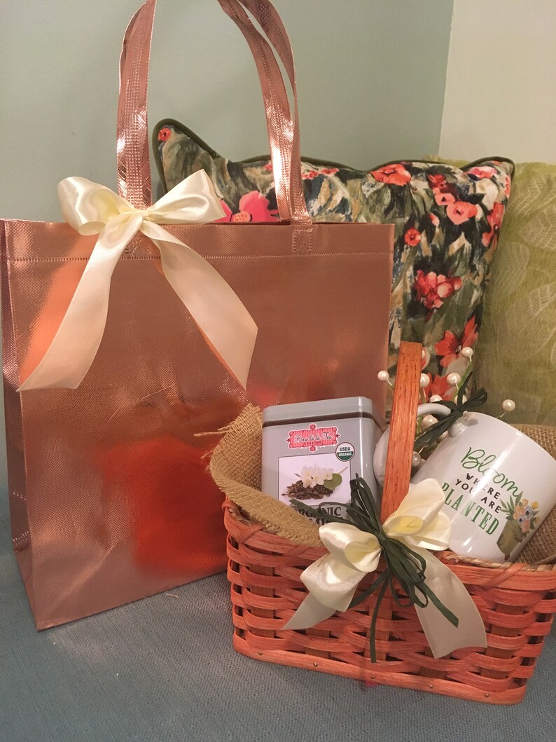May include: A gift basket with a rose gold tote bag and a small orange woven basket. The basket contains a tea tin, a mug with the text "Bloom where you are planted", and a burlap liner. Both baskets are tied with cream-colored bows.