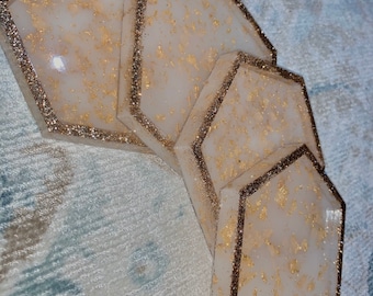May include: Set of four white hexagon-shaped coasters with gold glitter accents. The coasters are stacked on top of each other.