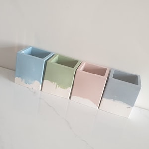 Peut inclure: Quatre petits pots carrés en béton de couleur bleu clair, vert, rose et gris avec un effet de goutte blanche en bas.