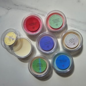 Könnte beinhalten: Eine Sammlung von zehn kleinen, durchsichtigen Glasbehältern mit Deckeln, die Wachs-Schmelzen enthalten. Die Behälter sind in einem Kreis auf einer weißen Marmorfläche angeordnet. Jeder Behälter hat ein farbiges Etikett mit dem Namen des Duftes und den Worten "The Original House Co."