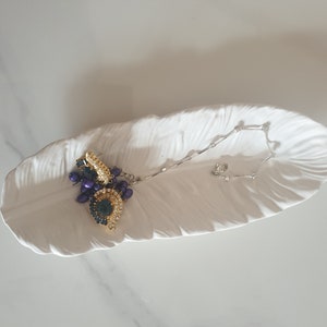 May include: A white ceramic feather-shaped dish with a gold and purple beaded necklace resting on it.