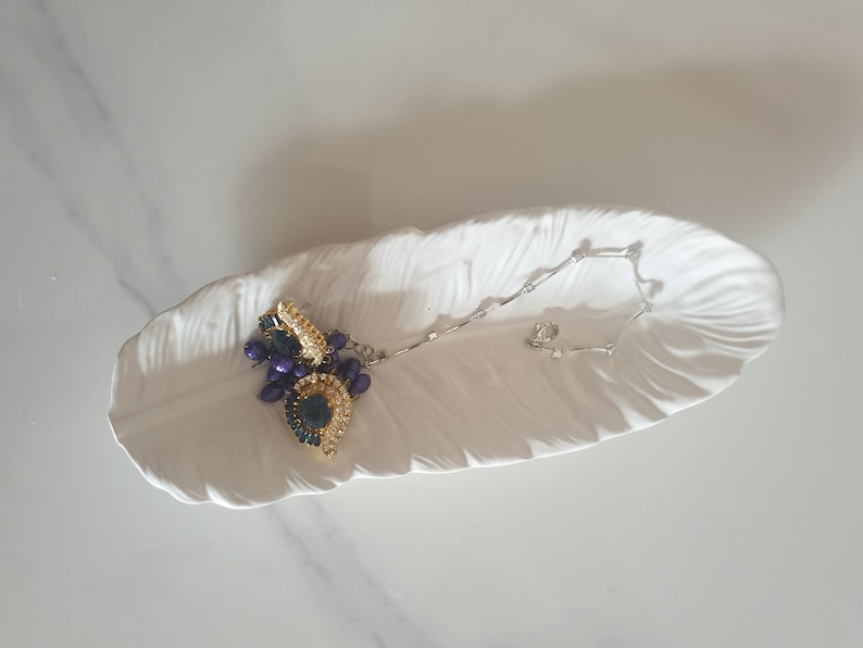 May include: A white ceramic feather-shaped dish with a gold and purple beaded necklace resting on it.