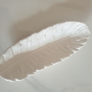 May include: White ceramic feather-shaped dish. The dish is designed to look like a real feather with detailed ridges and a smooth surface.