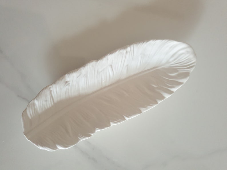 May include: White ceramic feather-shaped dish. The dish is designed to look like a real feather with detailed ridges and a smooth surface.