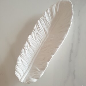 May include: White ceramic feather-shaped dish. The dish is designed to look like a real feather with detailed ridges and a smooth, glossy finish.